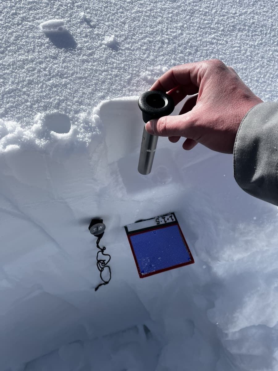 Stainless mini-coring tool held over the snowpit wall beside a crystal card and magnifier